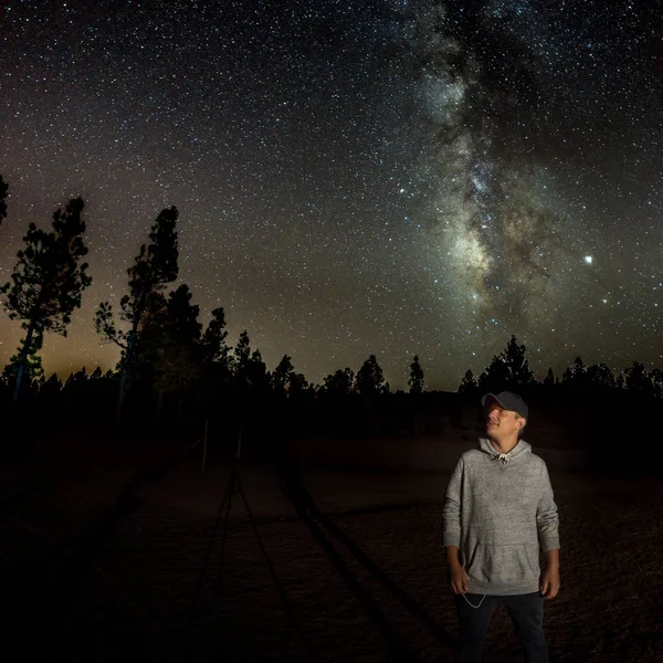 Ο νεαρός παρατηρεί έναστρο ουρανό. Βουνά, περιτριγυρισμένα από πευκοδάσος στο φόντο νυχτερινό τοπίο με πολύχρωμο Γαλαξία, αστέρια, πλανήτες και πεφταστέρι — Φωτογραφία Αρχείου