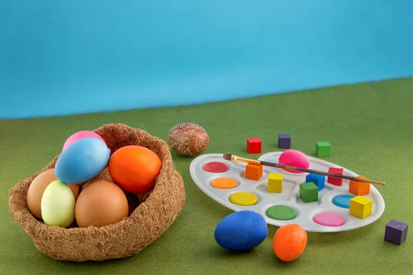 Colored easter eggs in the nest of straw. Palette with a brush for painting Easter eggs. Multi-colored cubes around. Green grass carpet and blue background.