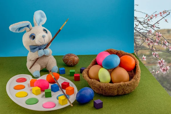 Colored easter eggs in the nest of straw. Gray plush rabbit with a palette with a brush paints Easter eggs. Multi-colored cubes around. Green grass carpet and blue background.