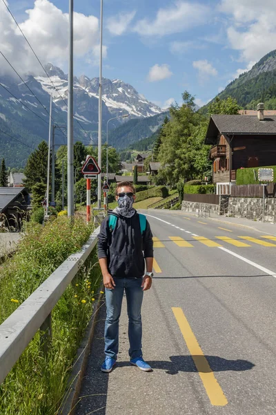 Genç adam boyun yürüyüşçüsü kullanıyor. Yüz maskesi gibi görünen braga. Sıcak güneşli bir gün, mavi gökyüzü ve beyaz bulutlar. Arka planda İsviçre Alpleri. Coronavirus koruma konsepti. Hava kirliliği kavramı.