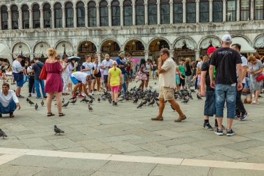 St. Mark Meydanı, VENICE, İtalya - 02 Ağustos 2019: sıcak güneşli bir yaz günü. Çok sayıda turist şehir güvercinleriyle fotoğraflanıyor. Çocuklar oyun oynar ve kuşlarla oynamaya çalışır..