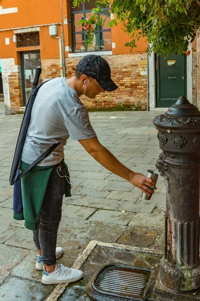 Metal termosu dolduran adam taze su için eski bronz sokak musluğunu kullanıyor. Geleneksel içme suyu çeşmesi Venedik İtalya.