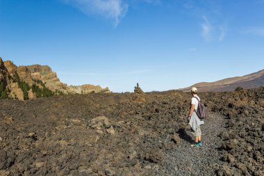 Sırtında sırt çantası olan şapkalı genç bir adam Teide Ulusal Parkı 'nda bir rota boyunca seyahat ediyor. Dağ yolunda yürüyüş. Etrafı lava kayaları ve bitki örtüsüyle çevrili. Tenerife.