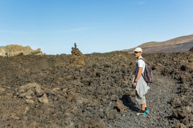 Sırtında sırt çantası olan şapkalı genç bir adam Teide Ulusal Parkı 'nda bir rota boyunca seyahat ediyor. Dağ yolunda yürüyüş. Etrafı lava kayaları ve bitki örtüsüyle çevrili. Tenerife.