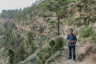 Sırt çantalı genç bir adam Tenerife 'nin batı yakasında bir rota boyunca seyahat ediyor. Dağ yolunda yürüyüş yapmak. Etrafı çam ağaçları ve lav kayaları ile çevrili. Kanarya Adaları.