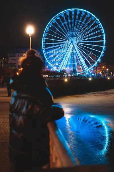 Mladá dívka na sobě ranní bunda sledování velké osvětlené ruské kolo v San Sebastian — Stock fotografie