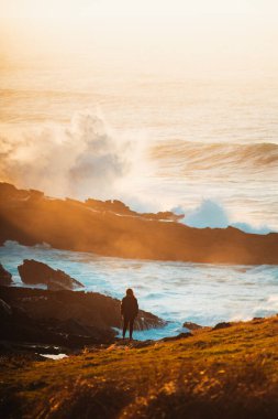 Yalnız kız büyük dalgaların kayalara çarpmasını izliyor.