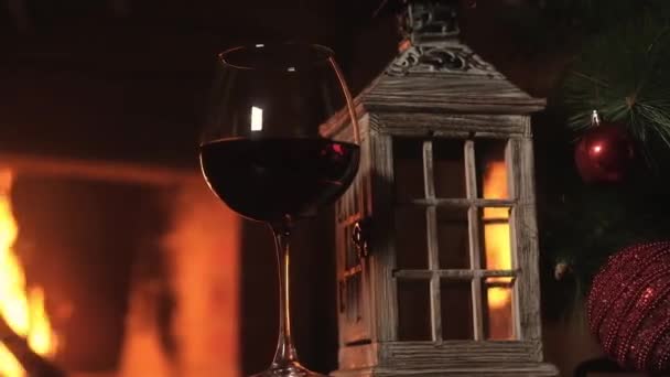 在壁炉前的桌子上 放着一杯红葡萄酒和木制灯笼烛台 与壁炉中的火势相对照 圣诞树上有装饰品 与朋友共度新年 — 图库视频影像