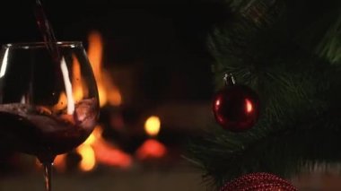 Kırmızı şarap, arka planda yanan Noel şöminesinin alevi üzerine saf şarap kadehinin içine dökülür. Noel yapay Noel ağacı ve güzel süslemeler. Aileyle yeni yıl tatili