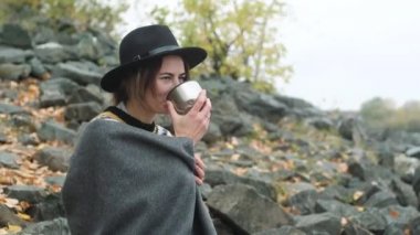beautiful girl in a black hat sits on a cool autumn day by the river Bank. He smiles and Drinks hot tea or coffee from a thermos. It is covered with a warm blanket and basks in nature. Close up.