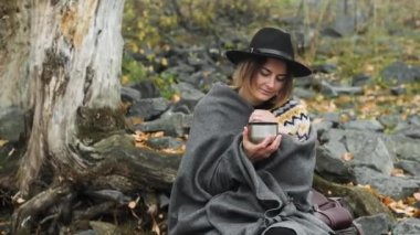Portrait of beautiful woman in black hat sitting on rocks on cool autumn day near riverBank smiles and Drinks hot tea or coffee from a thermos. It is covered with a warm blanket and basks in nature.