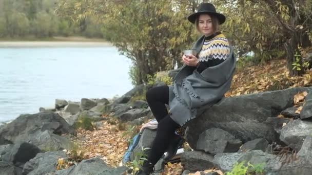 黒い帽子の若い美少女は川岸近くの大きな岩の上で涼しい秋の日に座っている 魔法瓶から熱いお茶やコーヒーを飲みます 毛布に包まれ 自然の中で日光浴 風景を見ると — ストック動画