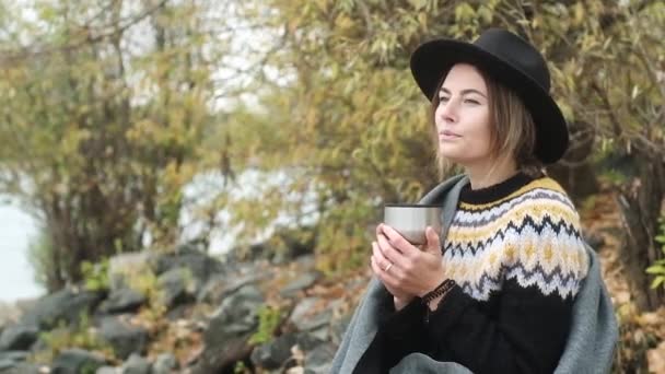 黒い帽子の若い美少女は川のほとりの涼しい秋の日に座っています 魔法瓶から熱いお茶やコーヒーを飲みます 毛布に包まれ 自然の中で日光浴 風景を見て 閉じろ — ストック動画