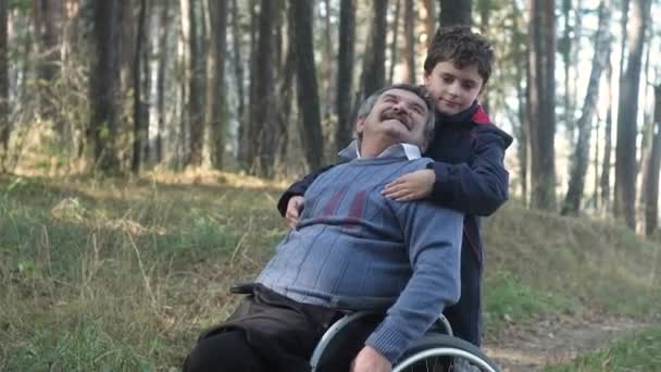 在一个阳光明媚的秋日 一个坐轮椅的老人和他的孙子在公园里散步 男孩拥抱着他的祖父 家庭护理 一个老弱病残 孙子在大自然中照顾他的祖父 — 图库视频影像