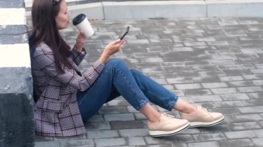 hipster girl in a casual jacket and jeans sits on the sidewalk near a concrete separation block and drinks their paper Cup of hot coffee or tea. The woman uses the phone reads the messages