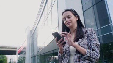 business woman in casual clothes with a modern phone in her hands stands on the background of the business center. A beautiful girl flips through the Internet or messages on her smartphone and smiles.