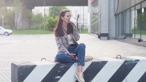 身着休闲装和牛仔裤的女孩坐在路上的混凝土隔板上 喝着热咖啡或纸杯茶 — 图库视频影像