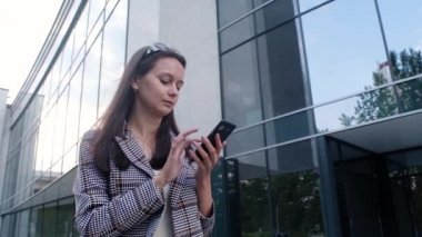 young girl in casual clothes with a modern phone in her hands stands on the background of the business center. Business woman typing a message on her smartphone. Communication by phone near the office