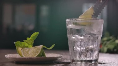 in kitchen on table is plate with sliced slices of fresh juicy lime and sprig of mint. The bartender picks up a piece of lime with tongs and puts it in a clear glass glass with ice cubes. slow motion.