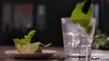 in kitchen on table is plate with sliced slices of fresh juicy lime and sprig of mint. bartender picks up a piece of mint with a pair of tongs and puts it in a clear glass with ice cubes. slow motion.