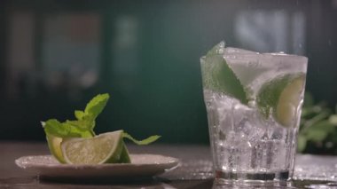 small clear glass glass with sparkling water and cubes of cold ice with slices of lime and a sprig of mint is on the table next to a plate of sliced lime. Citrus refreshing cocktail