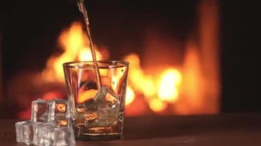 Sert viski boş şeffaf bardağa dökülür. Şöminedeki yanan bir alevin arka planında. Buz küplerinin yanına uzan. Panoramik video. Eğlenceli bir parti için güçlü bir alkol..
