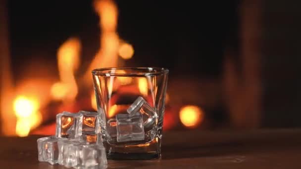 ウィスキーやブランデーやブランデーのための空のクリアガラスは暖炉の燃える火の背景にテーブルの上に立っています 氷のキューブの横に横たわっている 楽しいパーティーのための強いお祝いのアルコール — ストック動画