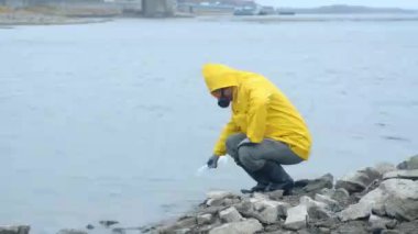 Sarı yağmurluk ve solunum maskesi takan ekolojist köprünün arka planıyla karşılaştırmak için nehirde su örneği alıyor. Temiz gezegen. Dünyayı umursuyorum. Nehirdeki suyun laboratuvar çalışmaları..