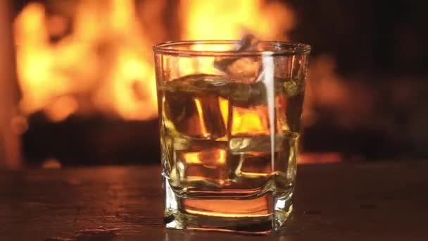 暖炉で炎を燃やすことを背景に古いウイスキーのガラスに落ちる氷のキューブのスローモーションのクローズアップ 酒のスプラッシュとドロップでパーティー 一晩中ごちそうだ 暖炉のそばで休んで — ストック動画
