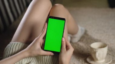 Kız yeşil ekranlı bir telefon kullanıyor. Kromakey çevrimiçi. Kazak giyen yetişkin bir kadın kanepeye oturur ve modern bir akıllı telefon tutar. Resim eklemek için yeşil maketi kullanın. Fotoğrafları parmaklarınızla kaydırın