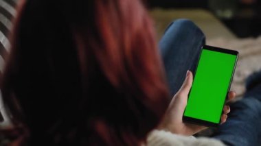 Kot pantolonlu kadın evinde kanepede dinleniyor ve elinde akıllı telefon var. Uygulamalar ya da resimler eklemek için yeşil model. Kız yeşil ekranlı telefonunu kullanarak fotoğraflara bakıyor. akıllı telefon kroma anahtarı