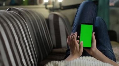 Genç kız evinde kanepede dinleniyor ve elinde yeşil ekranlı bir akıllı telefon tutuyor. Uygulamalar ya da resimler eklemek için yeşil model. İnternet bağımlılığı. Parmaklarınla hareket eden fotoğraflar.