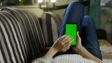 Genç kız evinde kanepede yatıyor ve yeşil ekranlı akıllı telefon kullanıyor. Uygulamalar ya da resimler eklemek için yeşil model. İnternet bağımlılığı. Elinde telefon olan kadın mesajı okuyor..