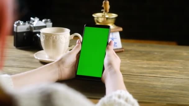 红头发的女孩坐在木制桌旁 拿着绿色屏幕的智能手机 桌上放着一杯咖啡 休闲的小玩意 上网成瘾 Chromakey Applications Programs — 图库视频影像