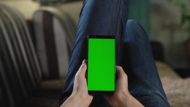 女孩躺在家里的沙发上 拿着装有绿色屏幕的智能手机 绿色模型以插入应用程序或图像 女孩用手指用手机翻看照片 智能手机着色键 — 图库视频影像