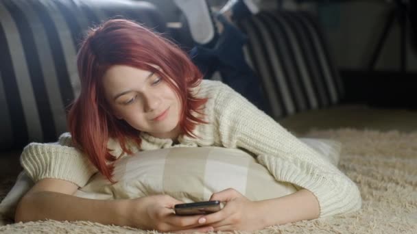 セーターに身を包んだ魅力的な女の子がスマホでソファに横になっています 電話でメッセージを読んでる女性 手のジェスチャーでニュースやメッセージをスクロールします 現代のガジェットで写真やビデオを表示 — ストック動画