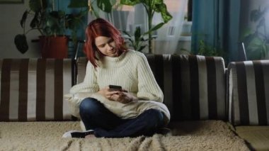 Akıllı telefonlu bir kız kanepede kazak ve kotla oturuyor. Çekici bir genç, iletişim kurmak ve mesaj yazmak için modern bir cep telefonu kullanır. İnternet bağımlılığı. Günlük hayatta cep telefonları.