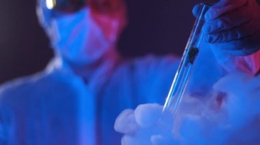 Laboratuvardaki mavi ışıklı, tulumlu bir araştırmacının elinde bir bitkiyle deney tüpü var. Araştırma için test örneği. Kriyojenik dondurucu. Sıvı nitrojen deneyleri.