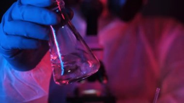 Neon ışıktaki bir laboratuvarda bir tıp bilimcisi veya kimyager şeffaf bir sıvıyı bir deney şişesinde sallıyor. Laboratuvar deneyleri neon mavisi kırmızı ışıkta. Laboratuvar ekipmanları. .