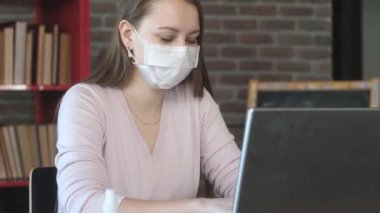 Maskeli genç kız dizüstü bilgisayarla çalışıyor. Merkez ofis. Karantina salgını sırasında izolasyon. Evde güvenlik işi. Bilgisayarda grip salgını. İş yerinde antiseptik.