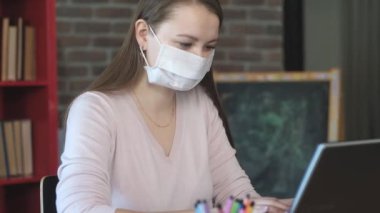 Beyaz tıbbi maskeli bir kız iş yerinde dizüstü bilgisayarla çalışıyor. Merkez ofis. Öğrenci ellerine antiseptik sıkar ve avuçlarını dezenfekte eder. İş yerinde güvenlik. İş yerinde antiseptik.