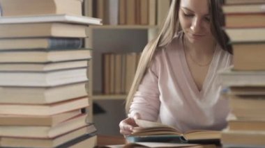 Kız kütüphanedeki masada birçok kitap ve ders kitabı arasında oturuyor. Öğrenci kitap okur ve sayfaları çevirir. Sınav hazırlığı. Öğrenci evde çalışıyor. Evde eğitim. Modern eğitim kavramı.