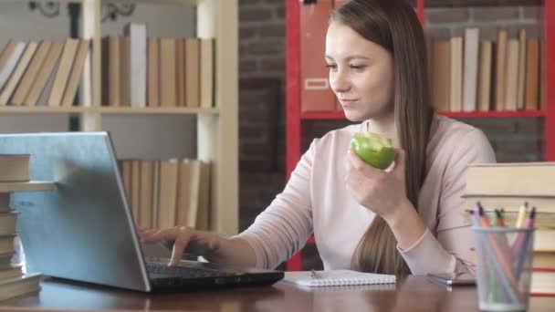 オフィスのノートパソコンで働いている美しい若い女の子 マネージャはジューシーな緑のAppleを噛み コンピュータを入力し続けます ホームビジネスの概念 プロのマネージャーはオフィスのコンピュータ上で動作します — ストック動画