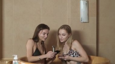 Mayo giymiş güzel, narin kız arkadaşlar Japon fıçısında sıcak suyla kaplıcada oturur ve telefonu eğlence için kullanırlar. Çekici kızlar deri ve vücut bakımı yapar..