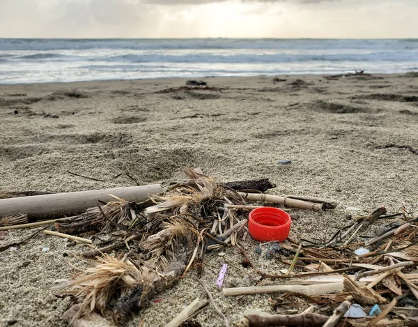 Kumlu deniz kıyısında kırmızı plastik şişe mantarı, kirlenmiş ekosistem, mikroplastik