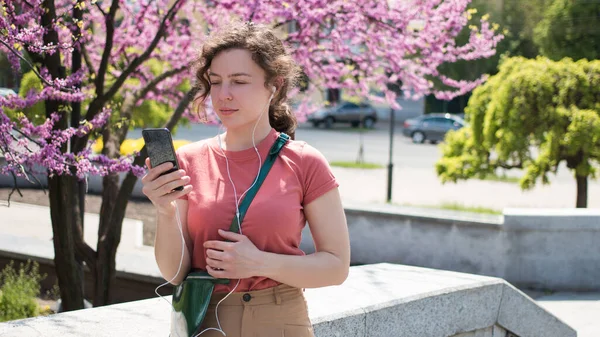 Pretty girl student listening audio cours, learning language, watching online webinar. Young woman wearing headphone using mobile phone for video call in city park outdoor,blooming tree