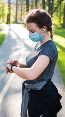 Tıp maskeli genç bir bayan parkta egzersiz yaparken müzik dinliyor ve akıllı saati ayarlıyor. Fitness kadını koşudan önce akıllı seğirme ayarlıyor. Covid-19 sırasındaki spor, karantina. 