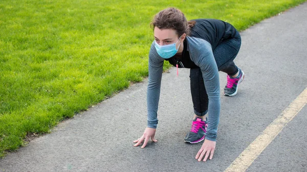 Sport during quarantine, coronavirus, covid-19 concept. Young beautiful sportive woman wearing face protective mask preparing to run outdoors in park. Outdoor run on athletics track in