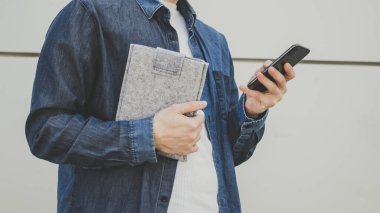 Beyaz tişörtlü ve kot pantolonlu bir adamın elinde akıllı telefonu gri duvarın arkasında tutarken. Okuma, okuma, izleme, giyme, e-posta kontrolü, kablosuz internet kullanarak sohbet. 