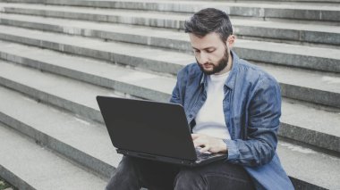 Dışarıda merdivenlerde dizüstü bilgisayar kullanan genç ve sakallı bir adam. Hipster milenyum erkeği kot yazısıyla yazı yazıyor, çalışıyor, blog yazıyor, internette sohbet ediyor, e-postaları kontrol ediyor. Öğrenci öğrenimi